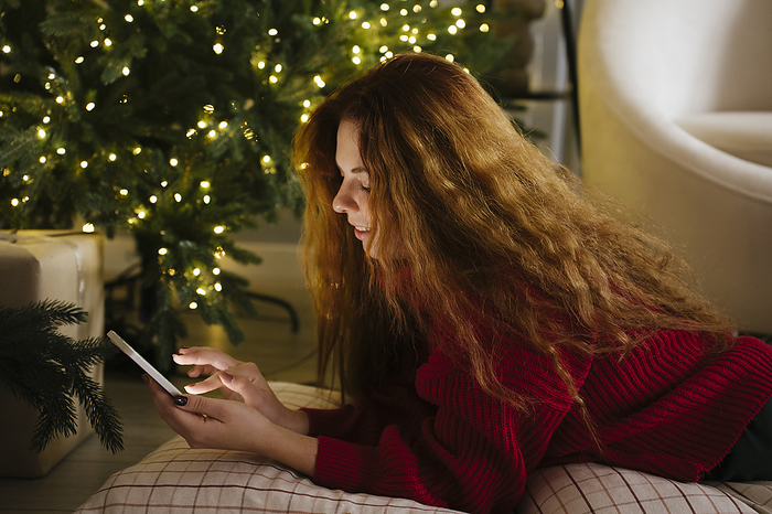 Smiling redhead woman using mobile phone near Christmas tree at home