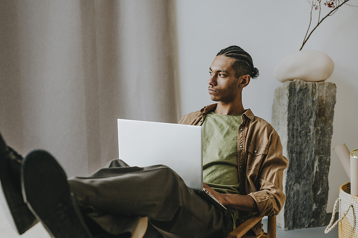 Thoughtful young man with laptop relaxing with feet on table at home