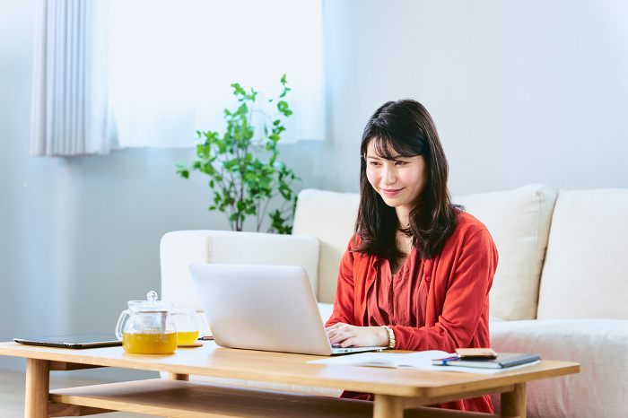 A smiling Japanese woman in her 30s teleworking and working from home with her computer spread out on a low table in front of the sofa in her living room.