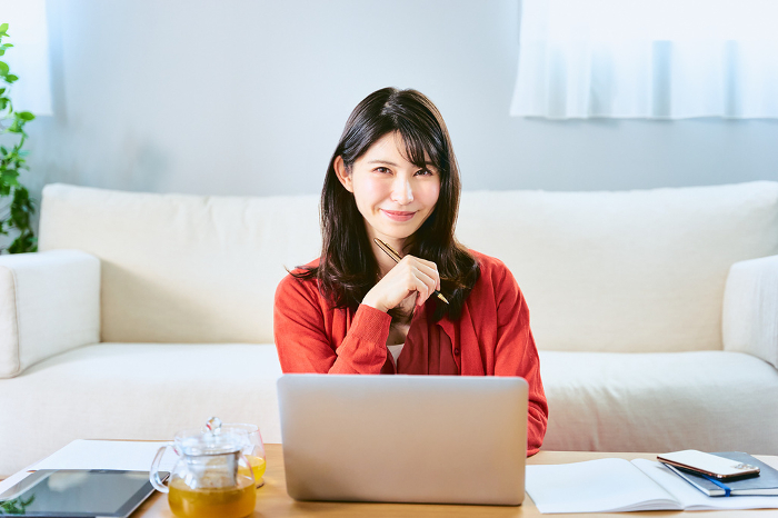 A smiling Japanese woman looking at the camera as she works or studies, holding a pen and spreading out materials, tablet or computer on a table in her living room (People)