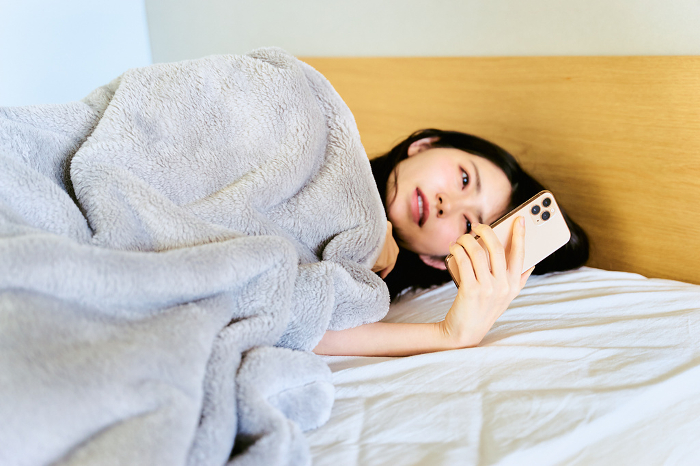 A Japanese woman in her 30s who overslept in her bedroom in the morning because of lack of sleep and poor health by lying on her bed and looking at her smartphone (People)