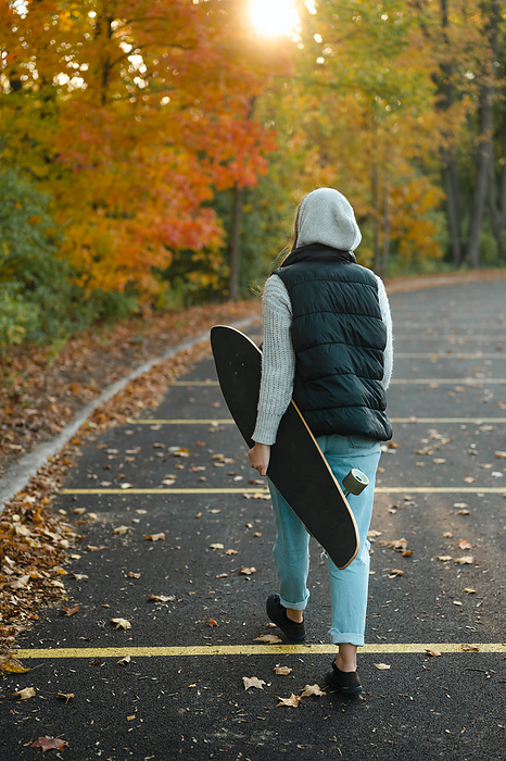 Woman walking with longboard in hand in parking lot with fall foliage, Chicago, Illinois, United States, by Cavan Images / Julia Maruyama