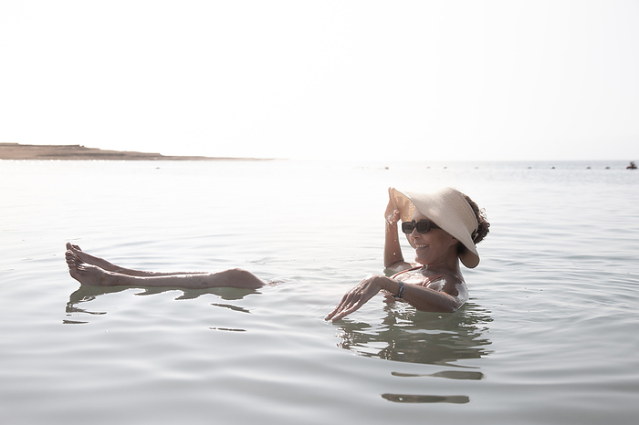 Woman wearing a hat and sunglasses, floating in the Dead Sea, Jordan, Madaba Governorate, Madaba, by Cavan Images / Marco Rof