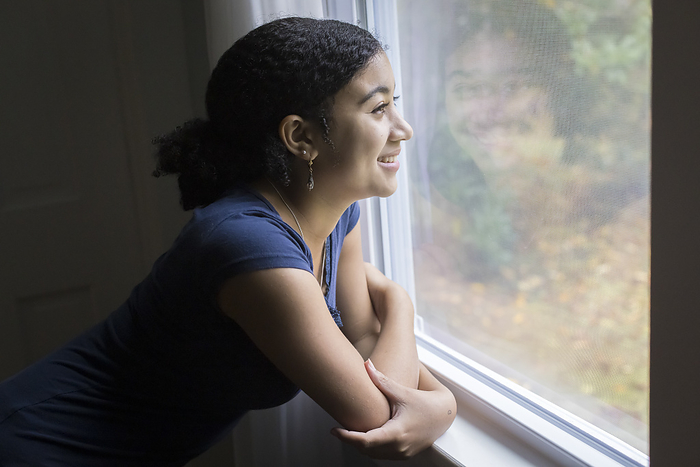 A smiling biracial teen girl looks out a window, Harwich, Massachusetts, United States, by Cavan Images / Julia Cumes