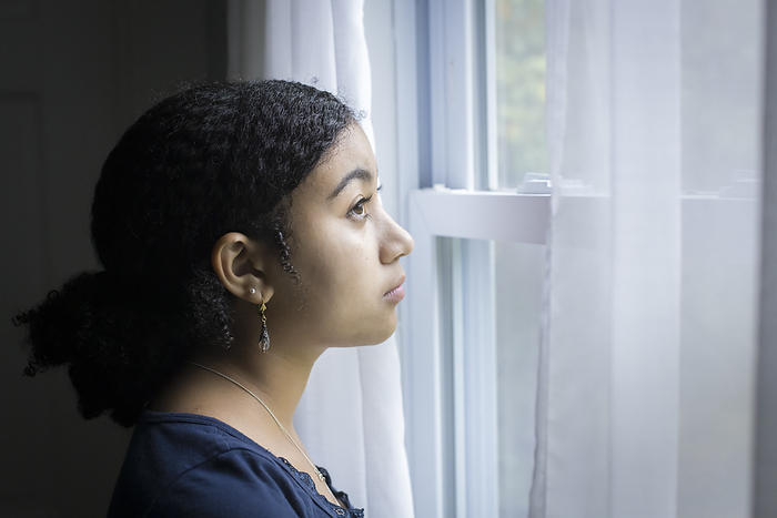 Biracial teen girl looks out a window with serious expression, Harwich, Massachusetts, United States, by Cavan Images / Julia Cumes