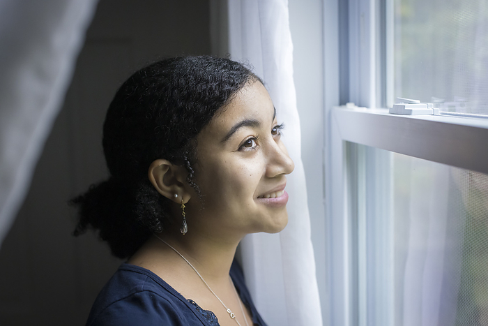 biracial teen girl looks out a window with a smile on her face, Harwich, Massachusetts, United States, by Cavan Images / Julia Cumes