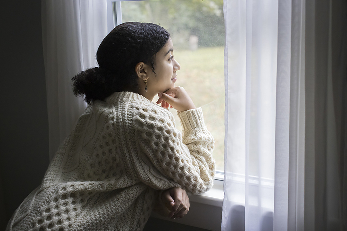 Biracial teen girl looks out a window with peaceful expression, Harwich, Massachusetts, United States, by Cavan Images / Julia Cumes