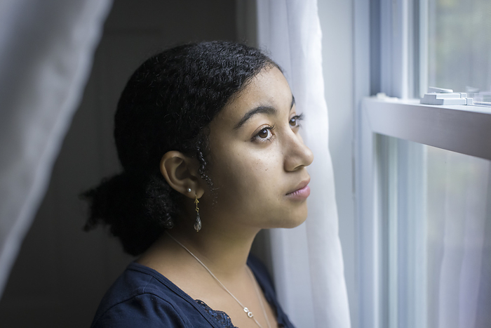 Biracial teen girl looks out a window with pensive expression, Harwich, Massachusetts, United States, by Cavan Images / Julia Cumes