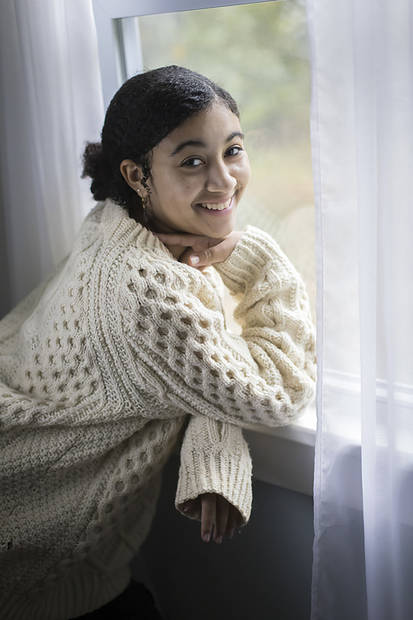 Smiling biracial teen girl at window looking at camera, Harwich, Massachusetts, United States, by Cavan Images / Julia Cumes