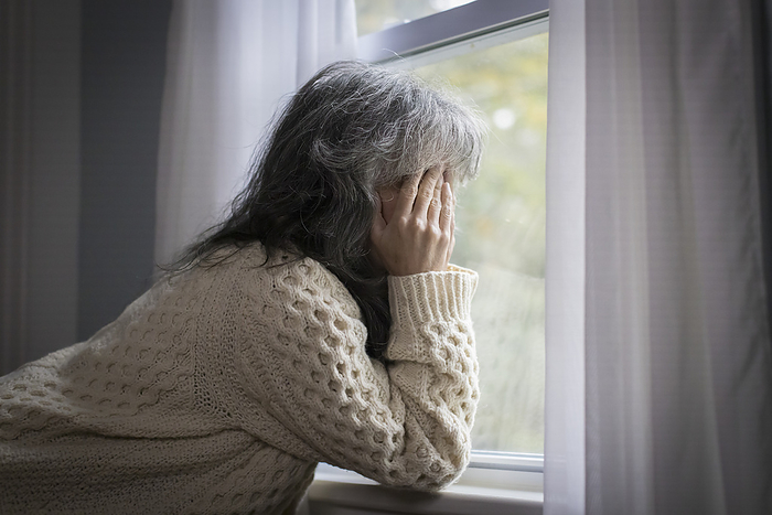 Older white woman by window with hands over face looking depressed, Harwich, Massachusetts, United States, by Cavan Images / Julia Cumes