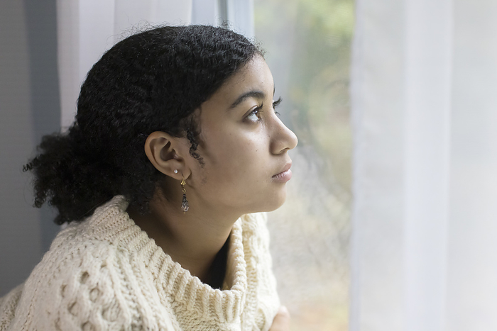 Biracial teen girl by window looking depressed, Harwich, Massachusetts, United States, by Cavan Images / Julia Cumes