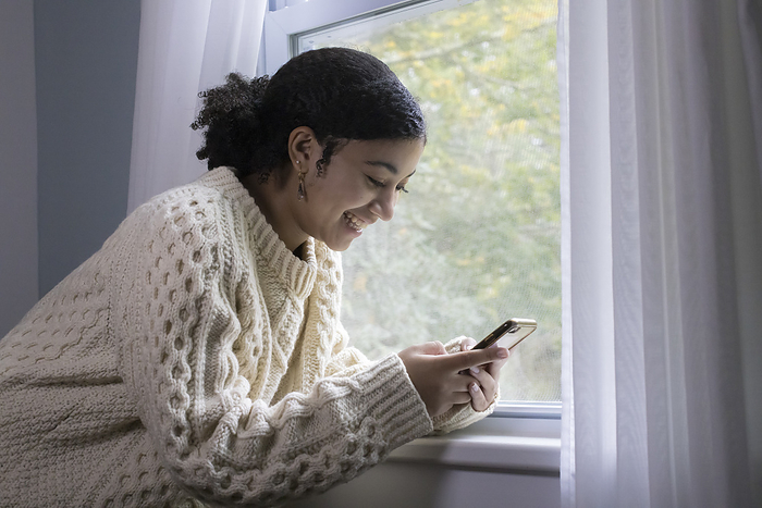 Biracial teen girl smiling and looking at her cell phone by window, Harwich, Massachusetts, United States, by Cavan Images / Julia Cumes