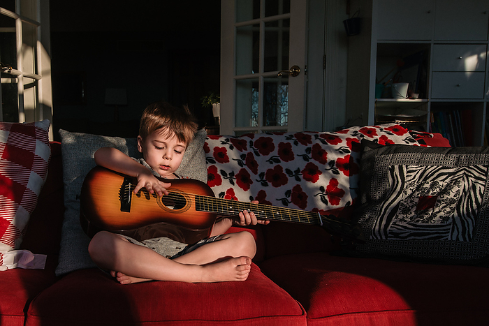 Preschool boy sits on couch and plays with child guitar in sunlight, Reading, Pennsylvania, United States, by Cavan Images / Liz DeGroff
