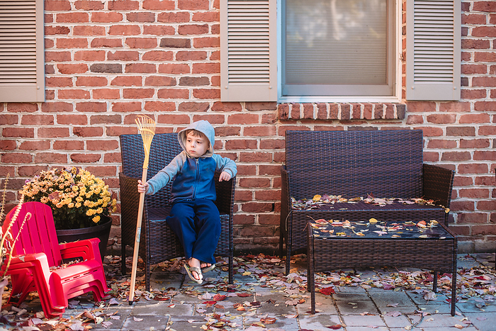 Preschooler in hoodie sits on patio holding small rake in the fall, Reading, Pennsylvania, United States, by Cavan Images / Liz DeGroff