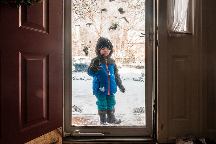 Preschooler in snow gear stands outside storm door on snowy day, Reading, Pennsylvania, United States, by Cavan Images / Liz DeGroff