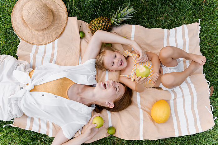 Mom and little daughter lie on a blanket, laugh and eat fruit, Milan, Lombardy, Italy, by Cavan Images / Liza Zavialova