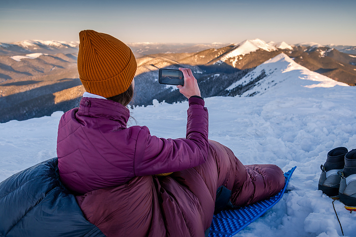 Woman in Sleeping Bag Using Mobile Phone Capturing The Snow Mountains, Bukovel, Ivano-Frankivsk Oblast, Ukraine, by Cavan Images / Artur Abramiv