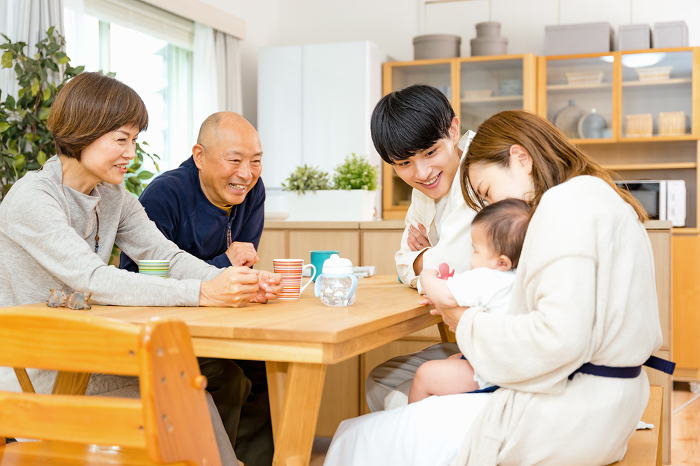 A Japanese family of grandfather and grandmother, sitting around a desk in the living room, delighted by their son and daughter-in-law's baby (People)