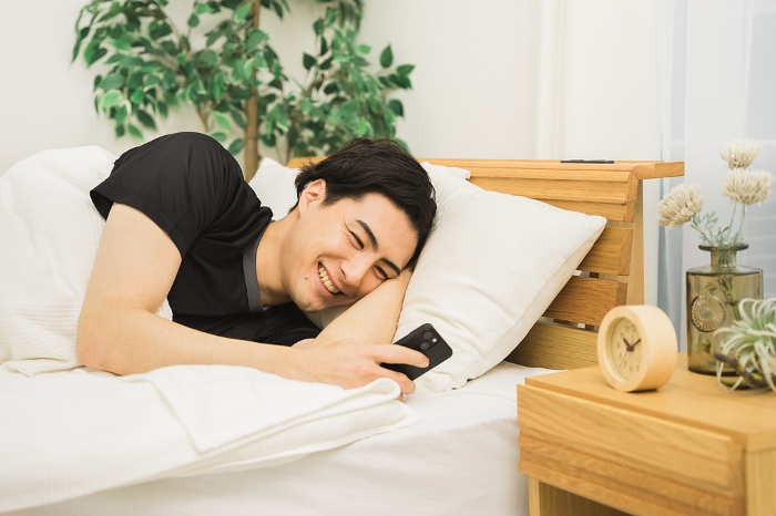 A Japanese man in his 20s or 30s operating a smartphone while lying on a bed in his room.