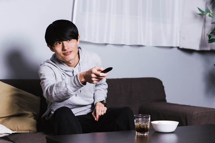 A Japanese man in his 30s operating a remote control to enjoy TV dramas and animations with drinks and snacks late at night in his room.