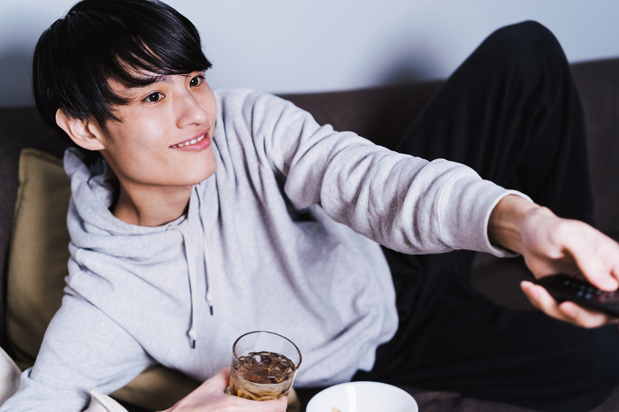 Lazy Japanese man in his 30s lying on the sofa, drinking and relaxing, operating the remote control and changing channels (People)