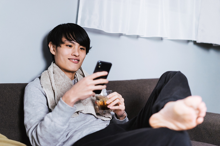 A Japanese man in his 30s with a towel wrapped around his neck after a bath, operating a smartphone with a glass in his hand.