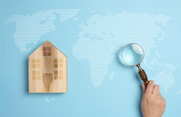 Wooden house on a world map background with a person s hand holding a magnifying glass, symbolizing the concept of searching for a country to move to Wooden house on a world map background with a person s hand holding a magnifying glass, symbolizing the concept of searching for a country to move to