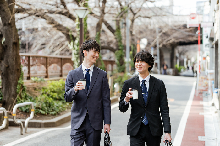Two Japanese male businessmen in suits commute to work holding takeout coffee in their hands while looking the other man in the face and conversing with him (People)