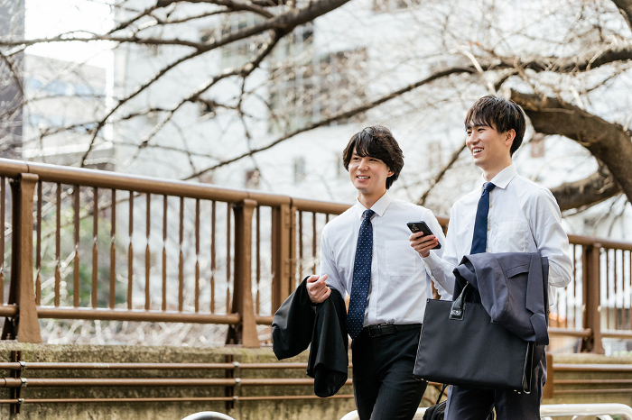 Two young, smiling, conversing Japanese men in suit shirts walking side by side on the greenway while searching and researching on their smart phones (People)