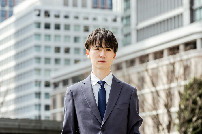Japanese man in a suit in his 20s looking seriously at the camera in an urban business district surrounded by buildings.