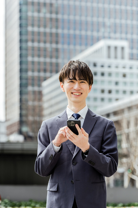 One Japanese man in his 20s, a businessman in a suit, looking at the camera, smiling and holding a smartphone in an office area.