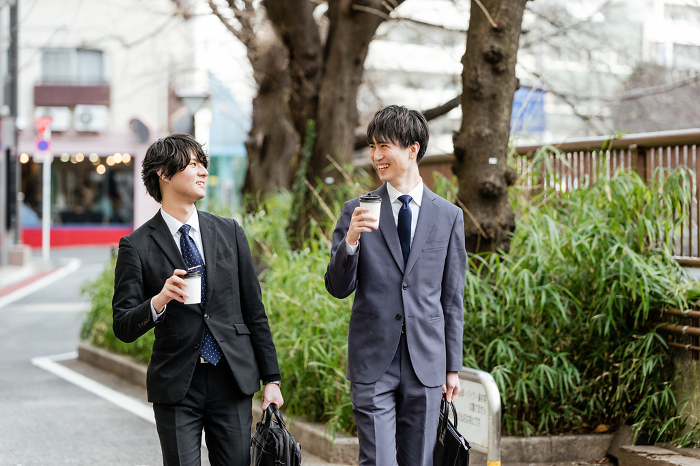 Two Japanese men in suits walking side by side with coffee in their hands, looking at each other and talking (People)