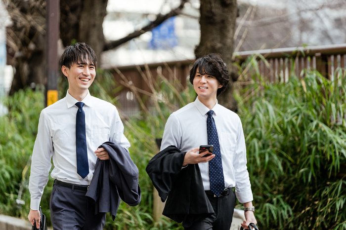 Two Japanese businessmen walking side by side with a colleague on the greenway, shirtless and without a suit, holding a smartphone in their hands (People)