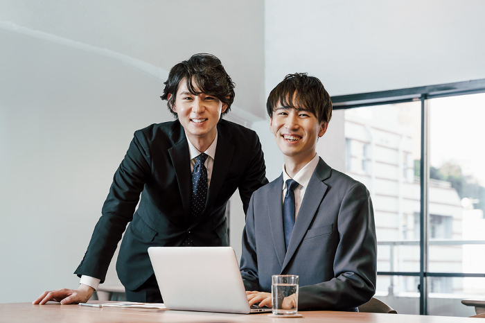 Two co-owners looking at the camera in an office conference room with laptop computers open and confident smiling faces (Male / Japanese / People)