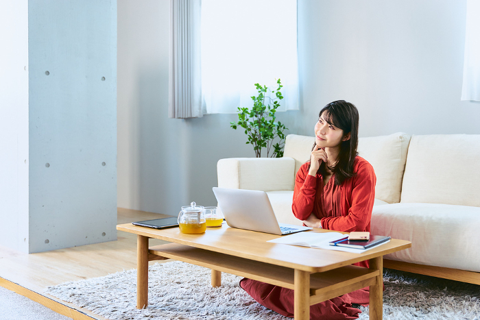 One Japanese woman with a laptop computer in front of her at her living room table, looking toward the copy space with a thoughtful expression (People)