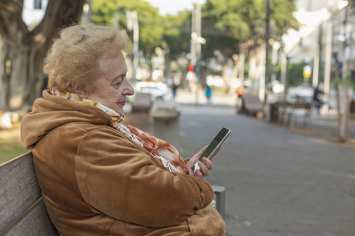 Senior woman sitting and using smart phone on city sidewalk bench