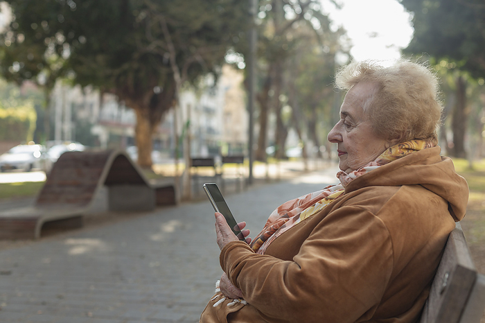 Senior woman sitting and using smart phone on park bench in city