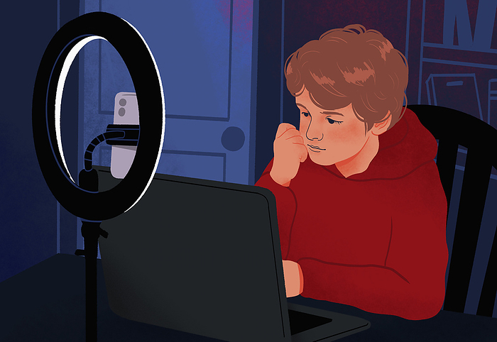 Boy using laptop in dark bedroom behind ring light and smart phone