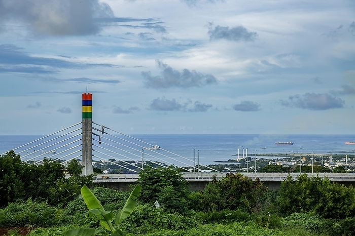 A highway with Mauritian emblem on pillar and a truck driving, surrounded by mountains and an overcast sky.A1-M1 Link Bridge At Grand River North West Valley on the island of Mauritius, by Doukdouk