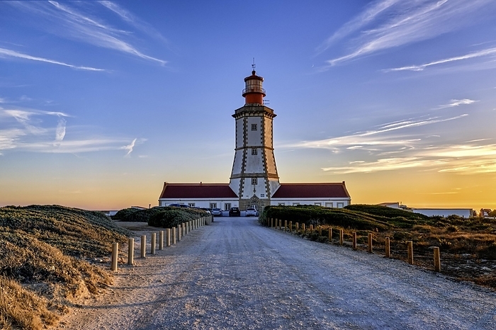 Beautiful of coastal lighthouse in warm colors of sunset, a clear path leading to it. Clear blue sky and some clouds, sun down over horizon, tranquil scene. Navigation beacon, safety for ships and small vessels, Atlantic ocean coast landscape, by Natallia Pershaj