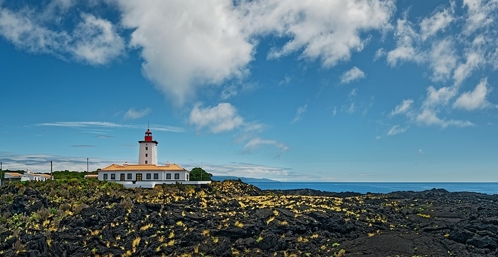 Wide lava flow with the striking lighthouse Farol da Ponta da Iha in front of a blue sky with white clouds, lava rocks coastal path Ponta da Iiha, Manhenha, west coast, Pico, Azores, by Michael Rucker