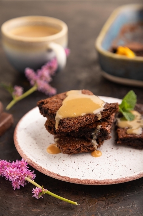 Chocolate brownie with caramel sauce with a cup of coffee on black concrete background. side view, close up, selective focus, by ULADZIMIR ZGURSKI