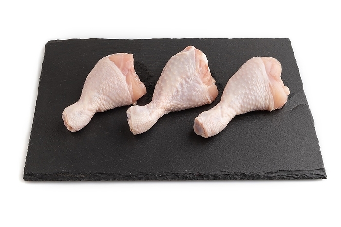 Raw chicken legs on a black slate cutting board isolated on white background. Side view, close up, by ULADZIMIR ZGURSKI