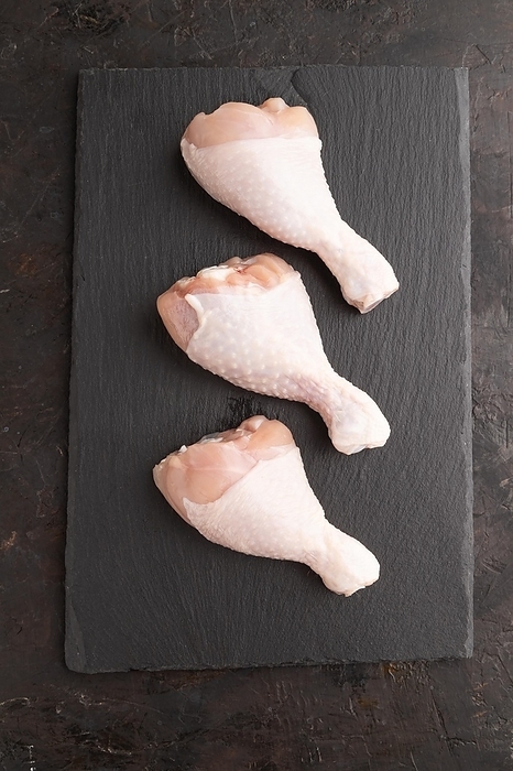 Raw chicken legs on a black slate cutting board on a black concrete background. Top view, flat lay, close up, by ULADZIMIR ZGURSKI