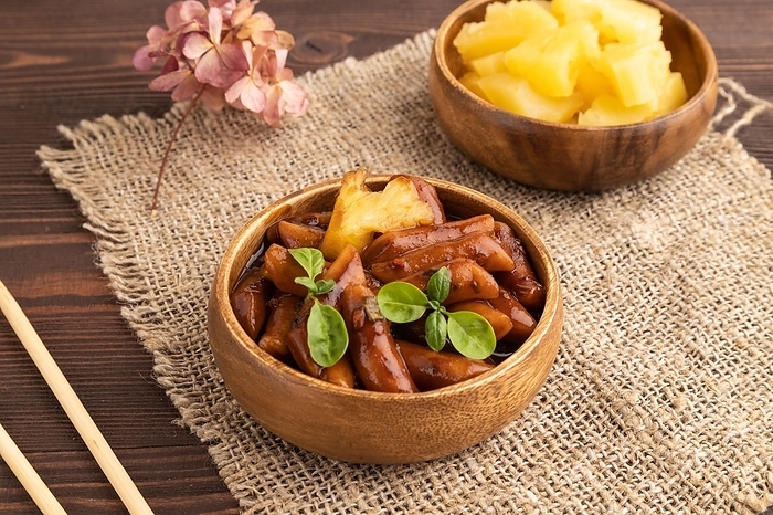 Tteokbokki or Topokki, fried rice cake stick, popular Korean street food with spicy jjajang sauce and pineapple on brown wooden background and linen textile. Side view, by ULADZIMIR ZGURSKI