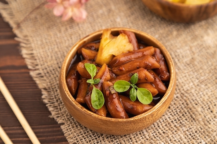 Tteokbokki or Topokki, fried rice cake stick, popular Korean street food with spicy jjajang sauce and pineapple on brown wooden background and linen textile. Side view, selective focus, by ULADZIMIR ZGURSKI