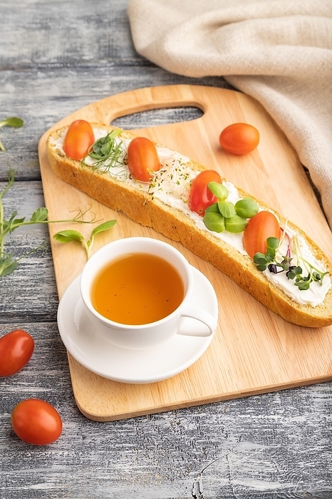 Long white bread sandwich with cream cheese, tomatoes and microgreen on gray wooden background and linen textile. side view, close up, by ULADZIMIR ZGURSKI