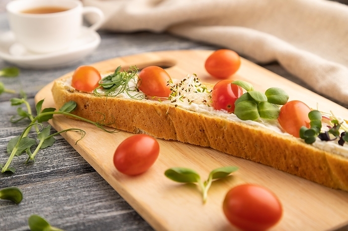 Long white bread sandwich with cream cheese, tomatoes and microgreen on gray wooden background and linen textile. side view, close up, selective focus, by ULADZIMIR ZGURSKI