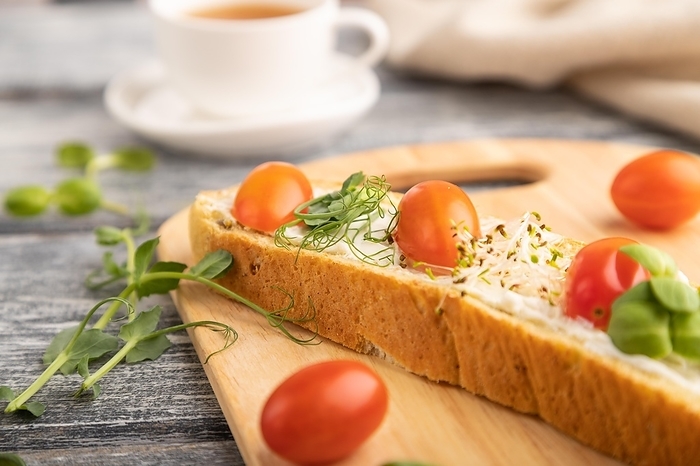 Long white bread sandwich with cream cheese, tomatoes and microgreen on gray wooden background and linen textile. side view, close up, selective focus, by ULADZIMIR ZGURSKI