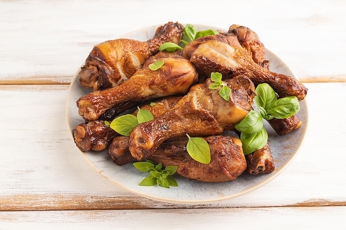 Smoked chicken legs with herbs and spices on a ceramic plate on a white wooden background. Side view, close up, by ULADZIMIR ZGURSKI